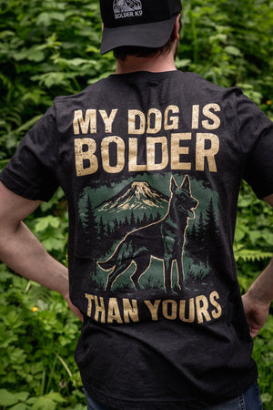 Man wearing 'My Dog Is Bolder Than Yours' t-shirt with mountain and dog graphic against greenery.
